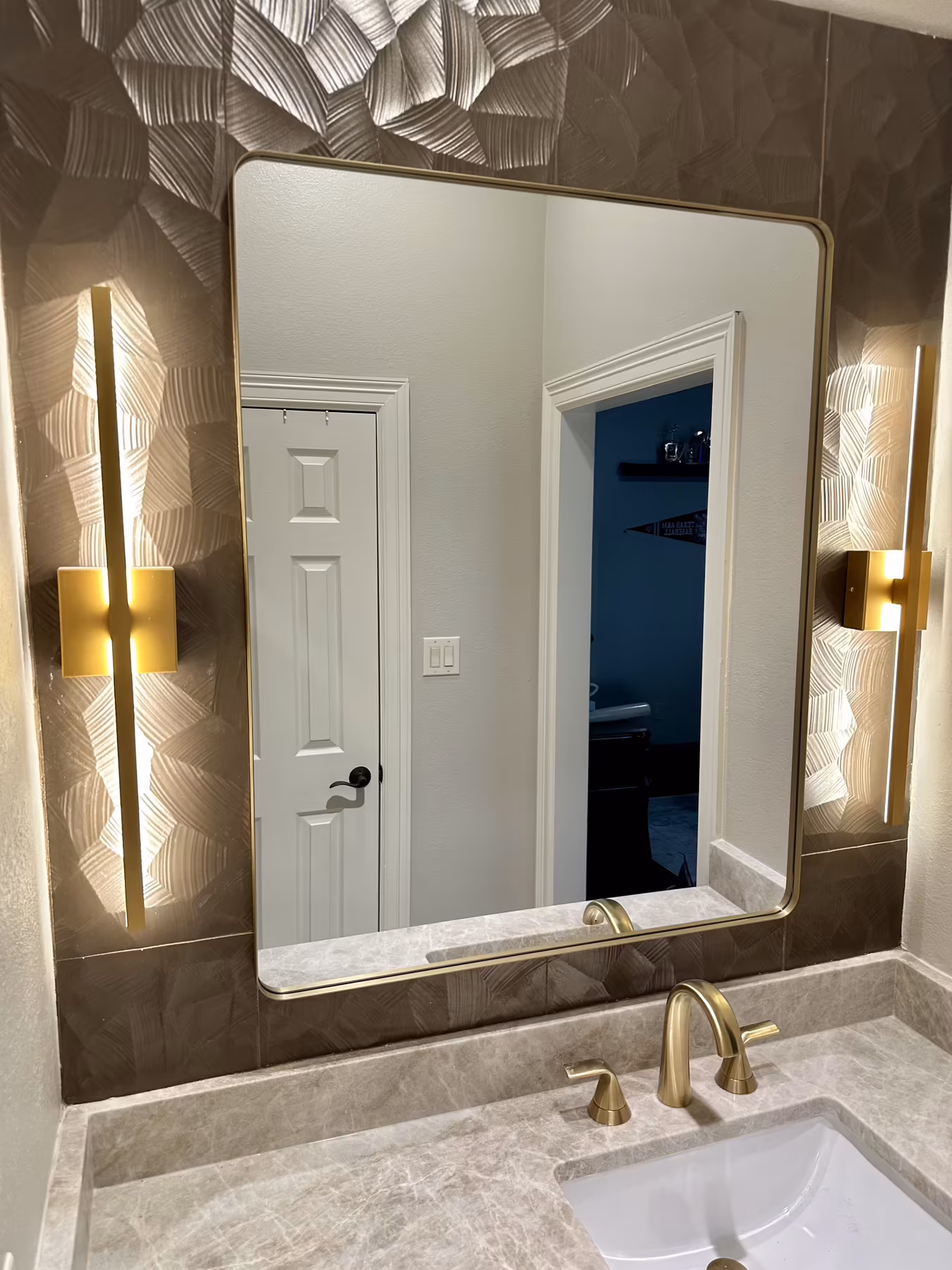 Gold-framed vanity mirror flanked by linear sconces on textured wallcovering